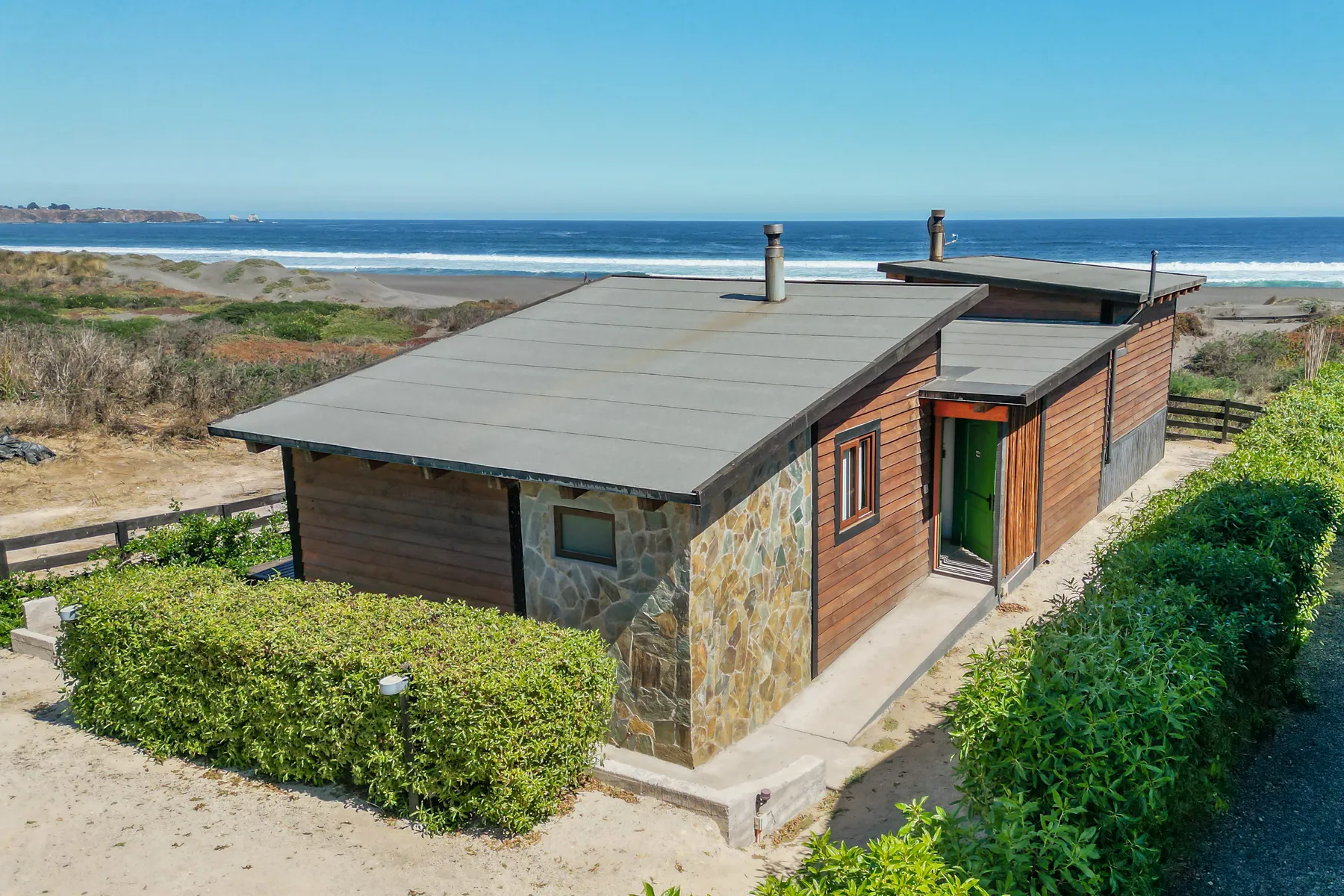 Cabaña Anakena vista desde afuera: volumen de madera y piedra al borde de las dunas, con la playa de Punta de Lobos detrás