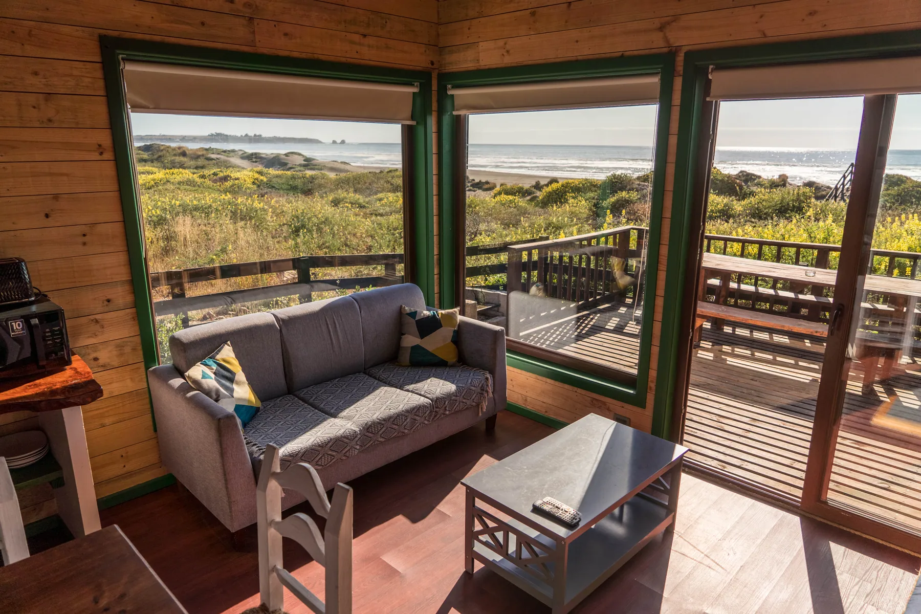 Living de la cabaña El Encanto con sofá y ventanales de esquina que dan a la terraza, con la playa al fondo