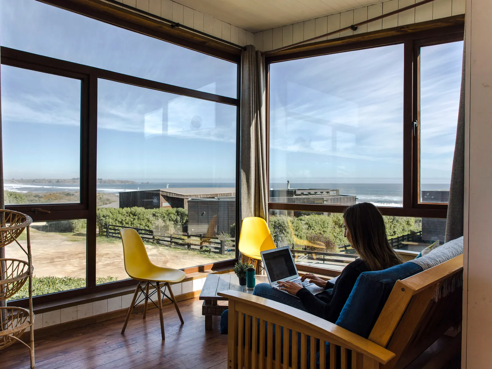 Mujer trabajando con su notebook en el living de una cabaña de Morros del Pacífico, frente al ventanal con vista al mar