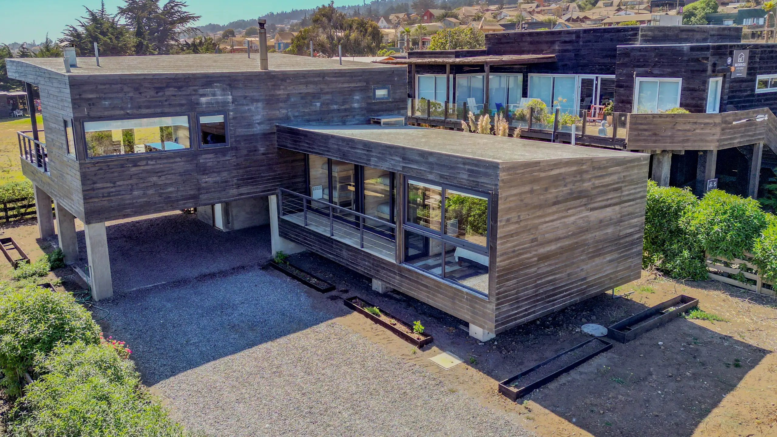 Vista aérea diurna de dos cabañas de madera dispuestas en L, con ventanales de piso a techo y senderos de maicillo entre los jardines